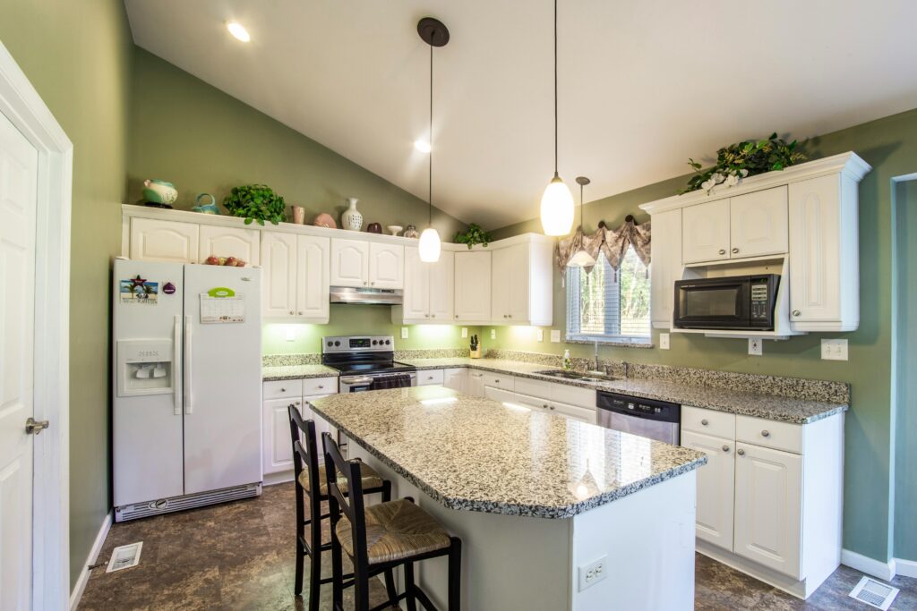 a photo of a kitchen with white cabinets, green walls and pendant lights over an island that are illuminated. A refrigerator and a microwave are visible