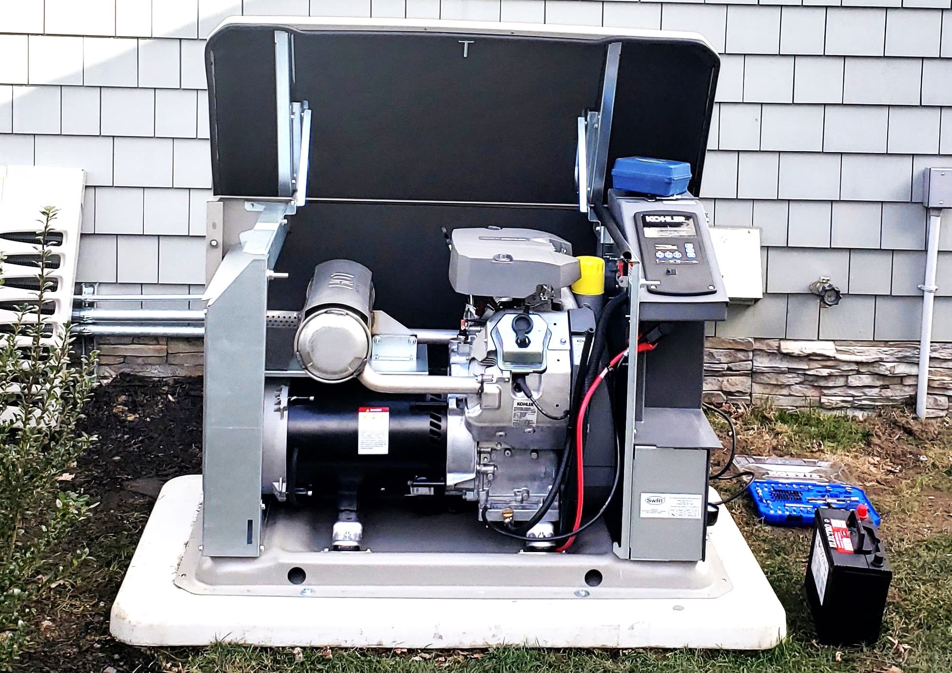 A photo of an opened kohler residential generator with a set of tools and testing equipment laying on the grass to the side of it. The side wall and foundation of a home are visible in the background.