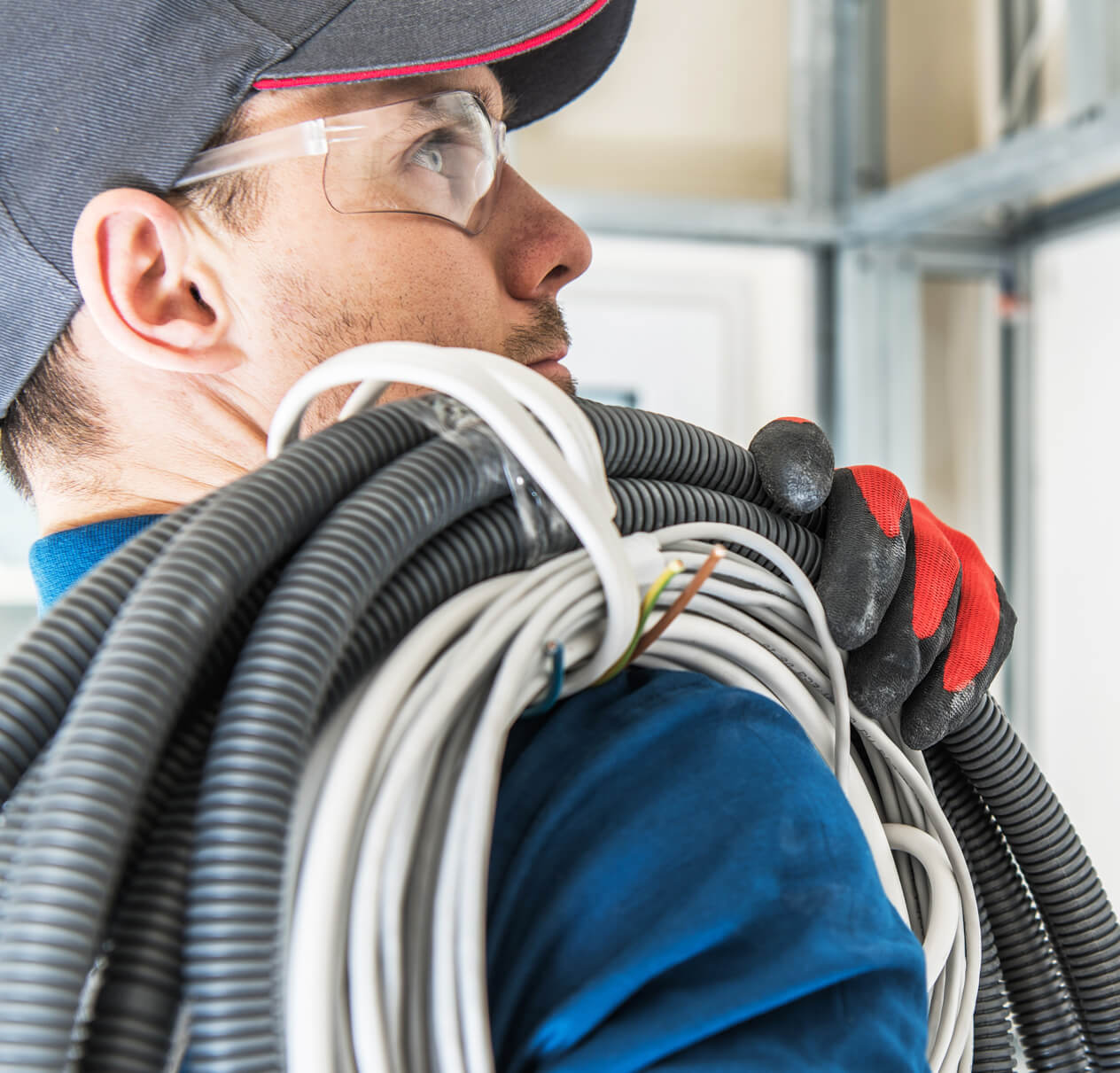 head and shoulders photo of electrical worker with cap, safety goggles and tubing and wiring coils wrapped over his shoulder.